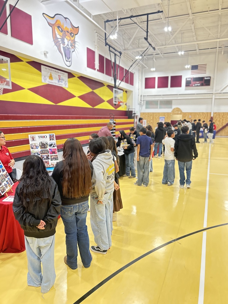 People stand in line at a school gym with a basketball court, yellow floor, and maroon walls featuring a tiger mascot.