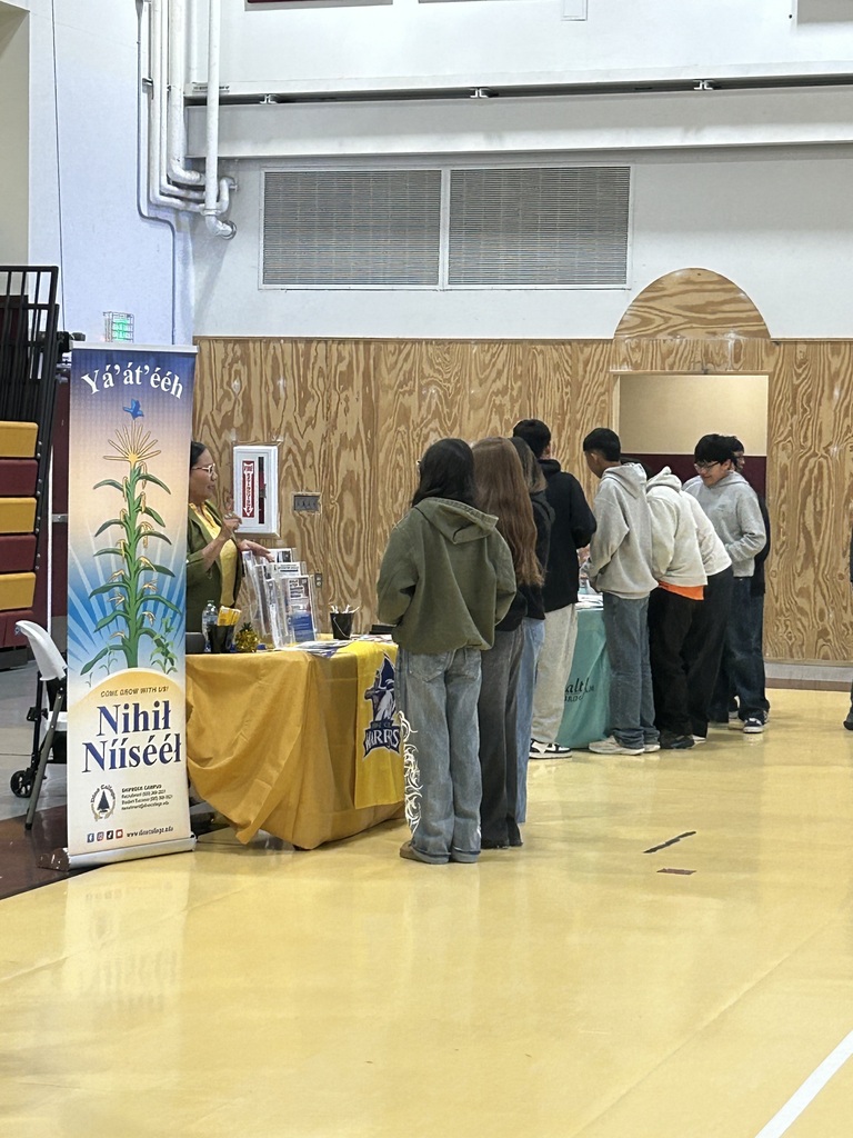People stand in line at an indoor booth with a yellow tablecloth and a sign with a plant illustration.