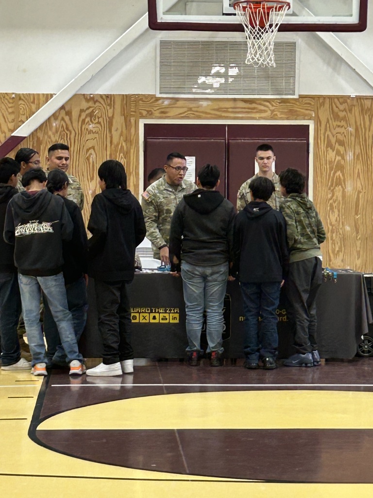 People gather around a table with a banner inside a gymnasium. One person is speaking to the group.