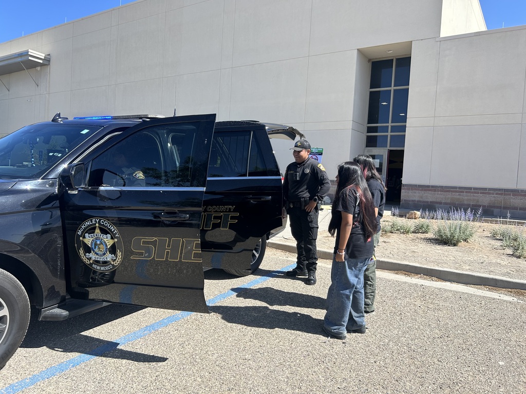 A law enforcement officer stands by a parked black SUV in a parking lot, with people nearby.