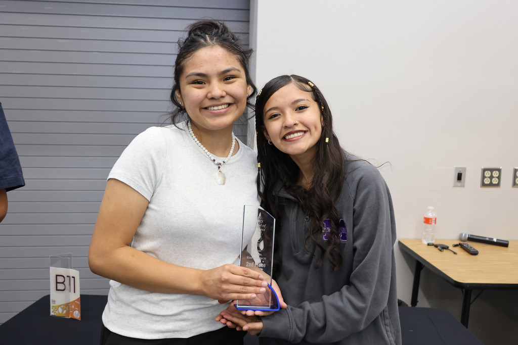 Two women, one holding an award, stand smiling with a backdrop of a white wall and table.