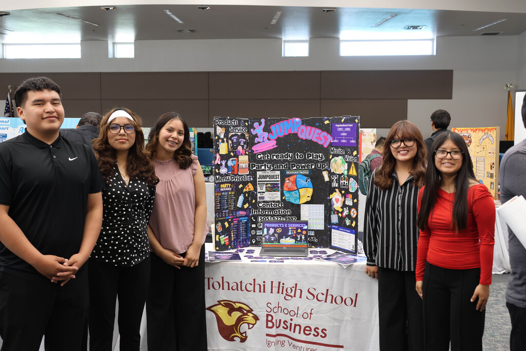 Four individuals pose at a table with a sign reading "Tohatchi High School School of Business."