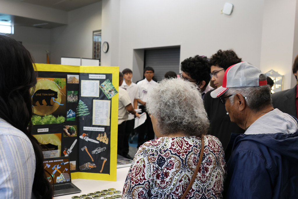 A group of people examines a display of photos and drawings on a board at a school event.