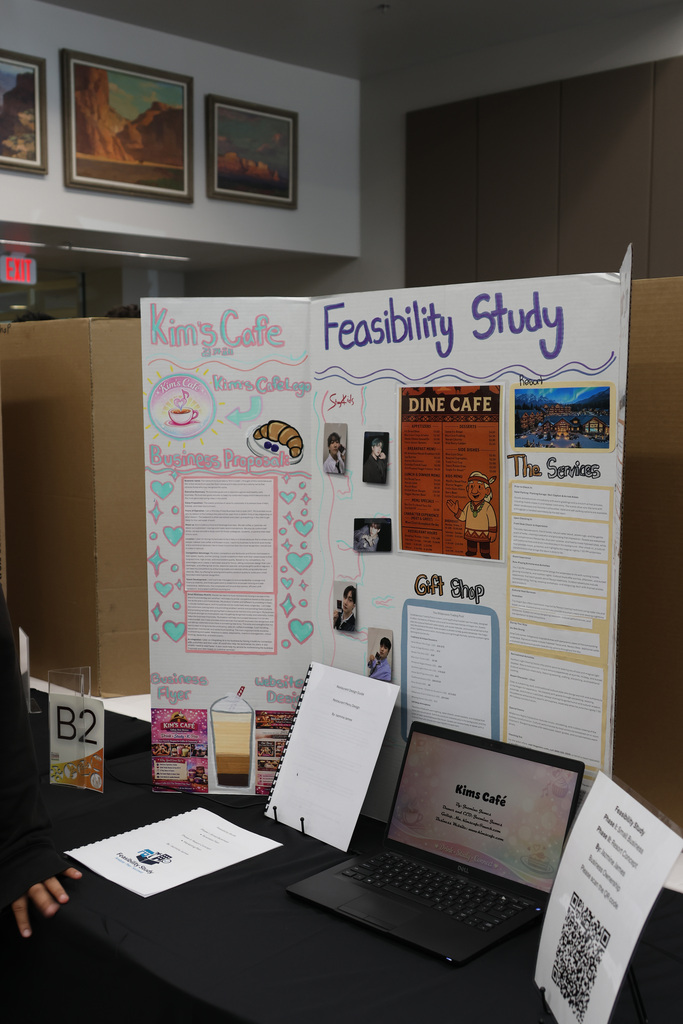 A table with a laptop, papers, and a QR code. Behind it, a poster titled "Feasibility Study" displays business concepts.