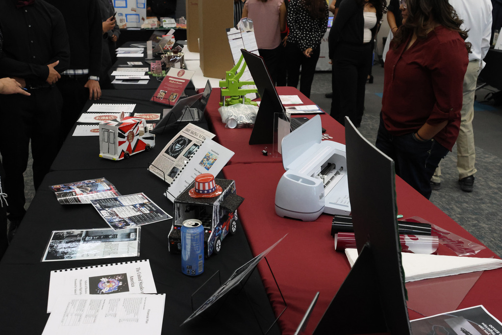 People stand around a table displaying a variety of items, including books, a can, and a printer.