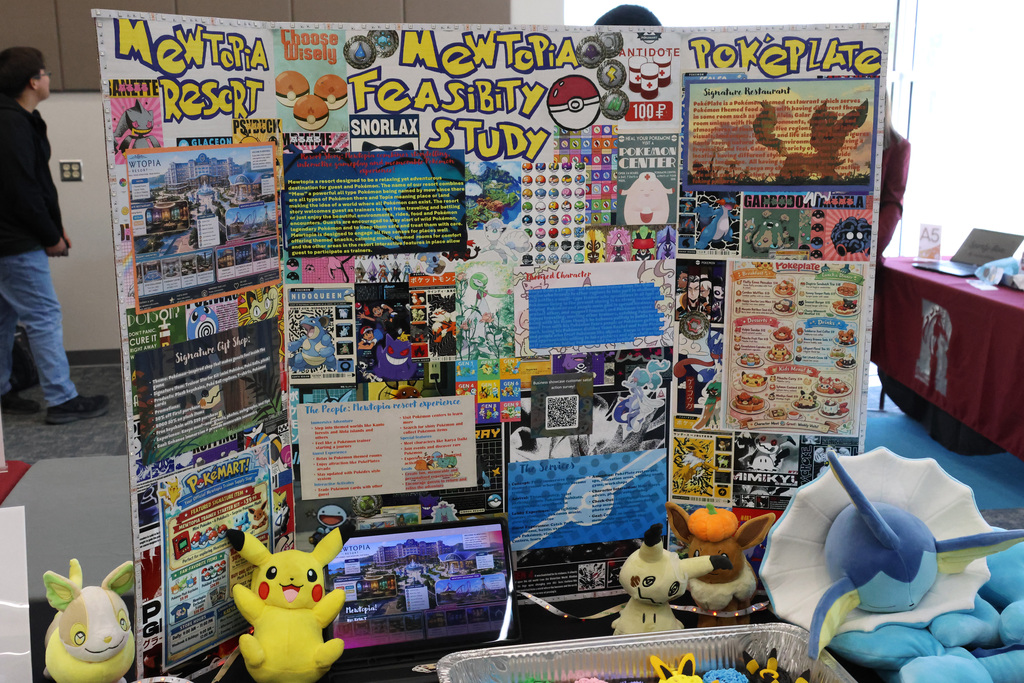 Display of a project board with images and text. A laptop, Pikachu plush, and other toys on a table.