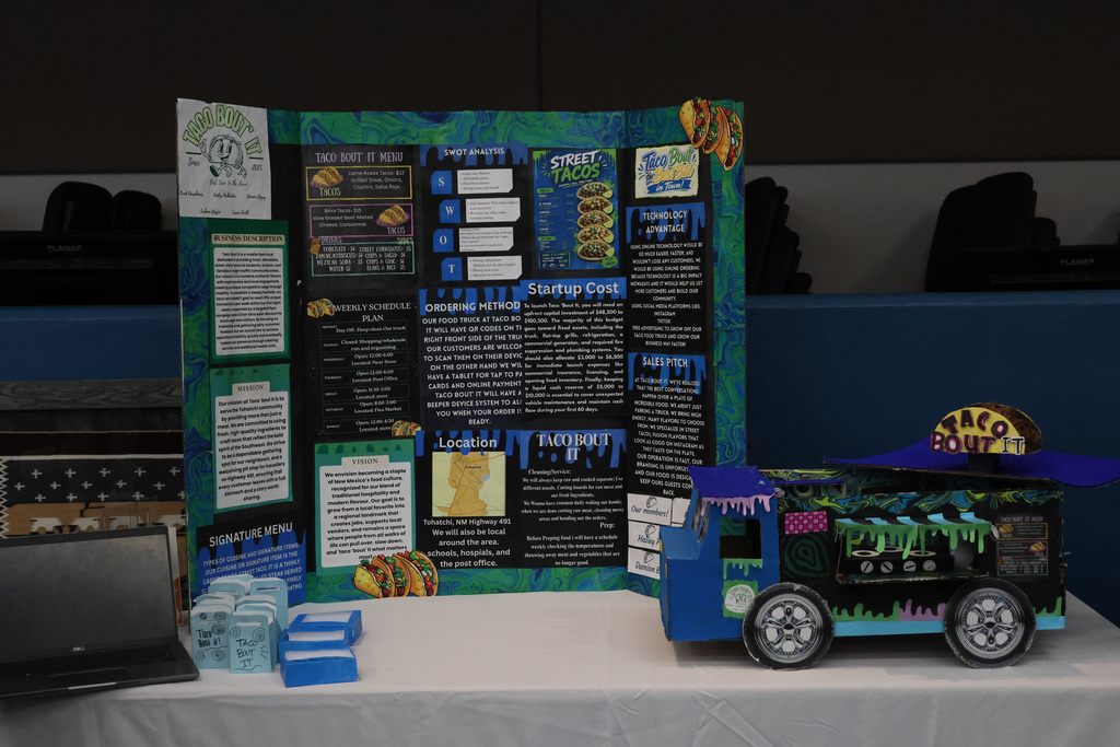 Display on a white table, featuring a blue and green poster with text and images, a model food truck with green and blue decorations.