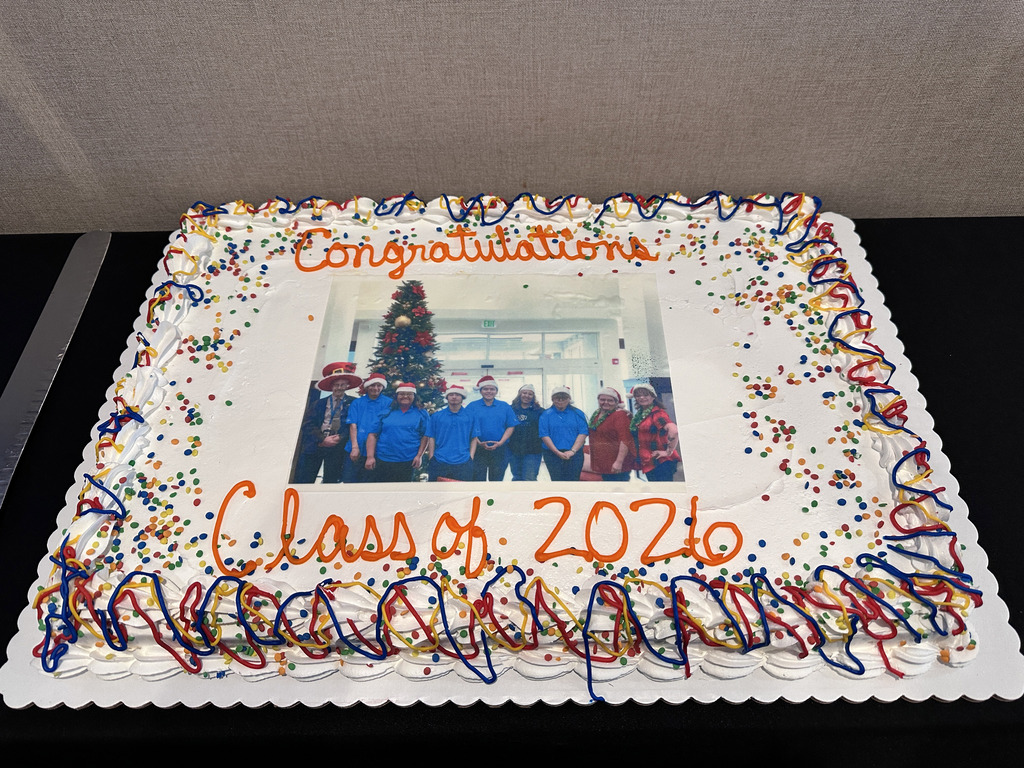 A square cake with colorful confetti. It features a photo of people in blue shirts with a Christmas tree in the background.