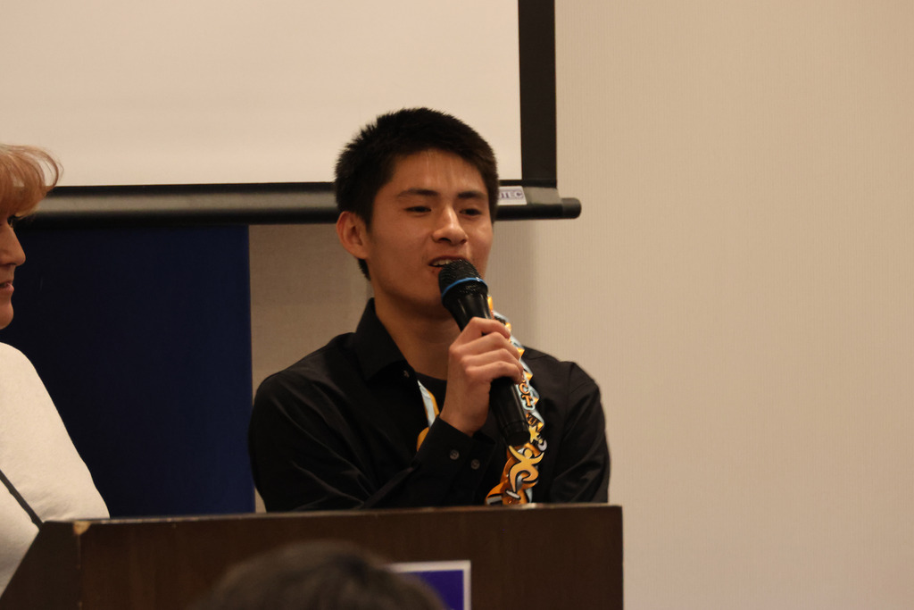 A person at a podium holds a microphone and speaks while another person stands nearby.