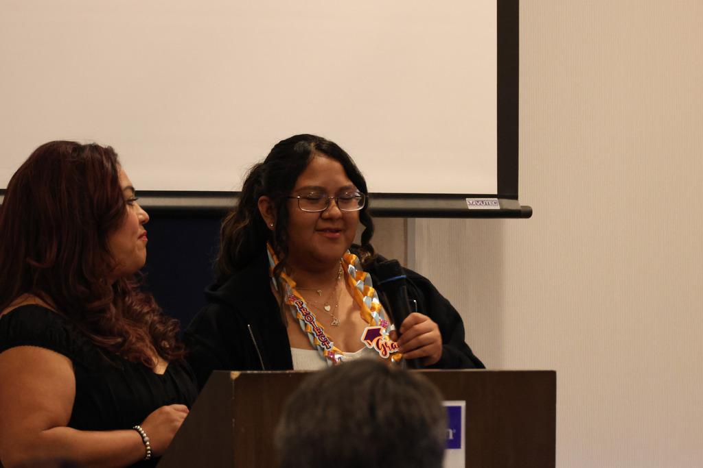 Two women at a podium. One woman speaks into a microphone, the other watches. Behind them, a projector screen is visible.