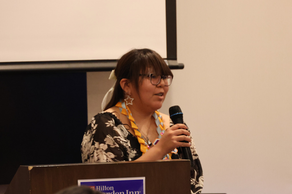 A woman wearing glasses and a floral shirt speaks into a microphone at a podium, with a Hilton banner behind her.