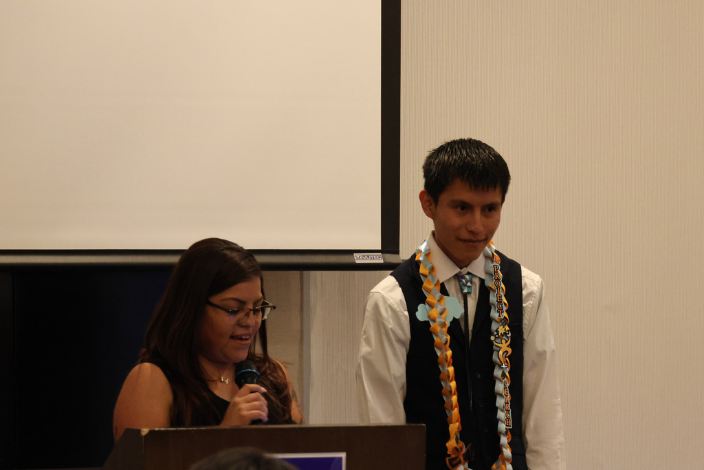 A woman with glasses speaks into a microphone at a podium. Beside her stands a man wearing a lei.