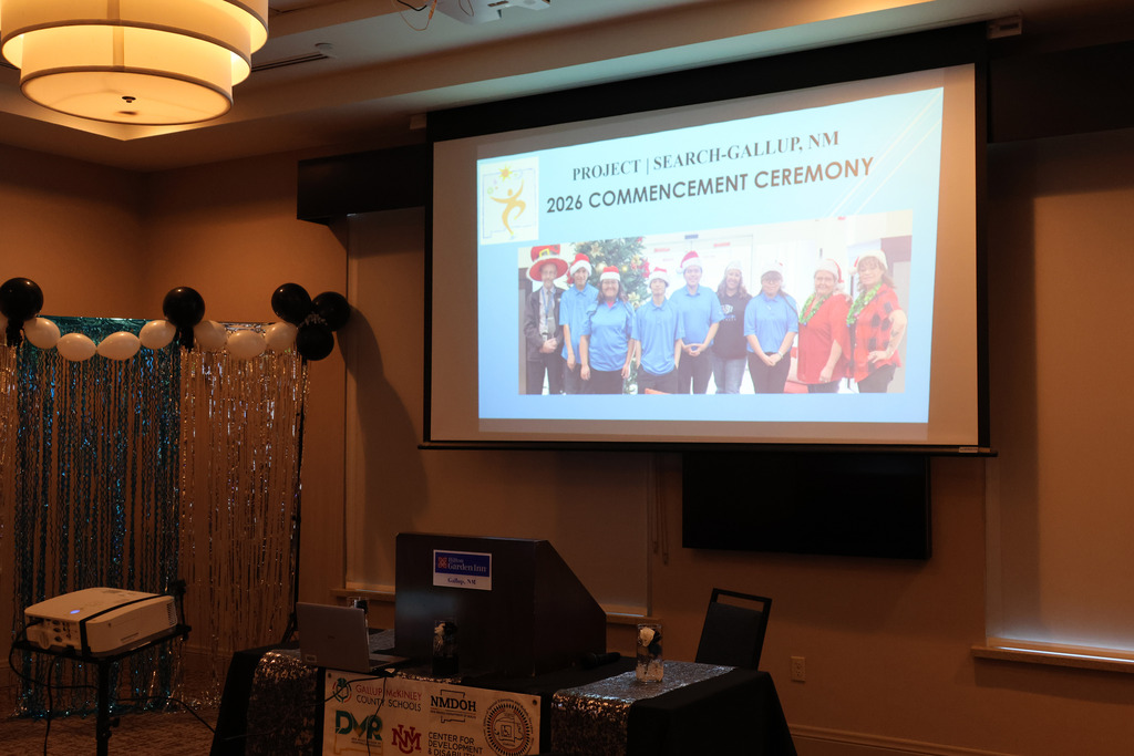 A presentation on a projector screen shows a group in matching blue shirts. A podium with a computer, chair, and projector is in the foreground.