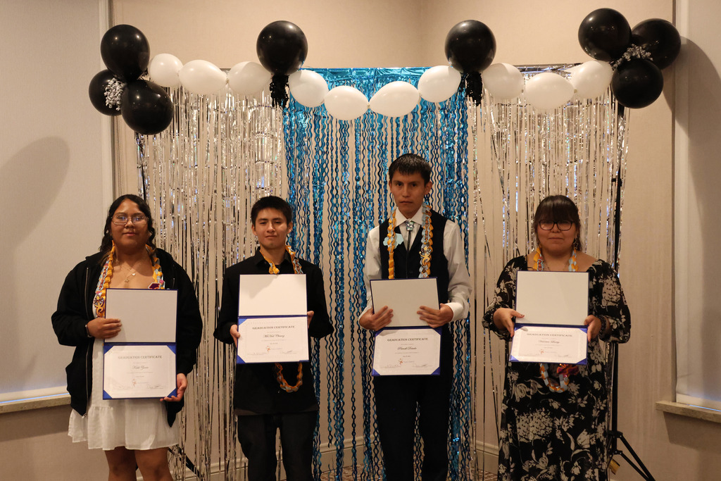 Four people stand with certificates in hand in front of a backdrop of silver tinsel and balloons.