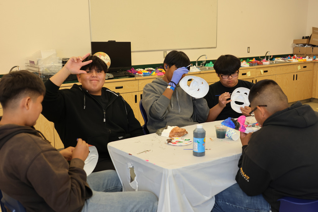 Five young men seated around a table. Three wear face masks, one with a painted smile. They appear to be working on art projects.