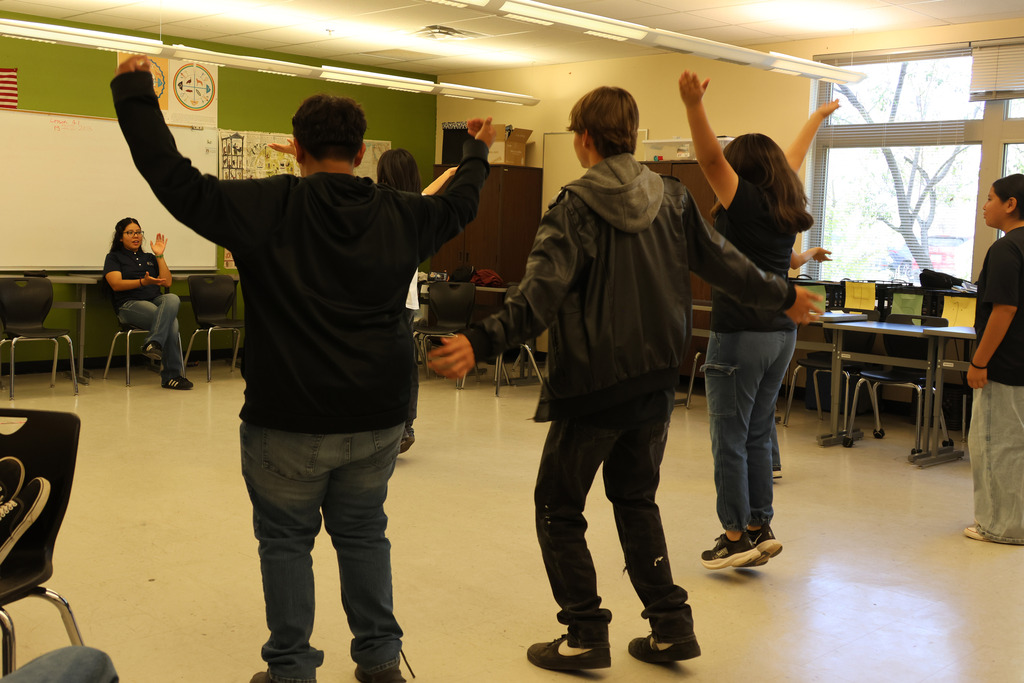 Several individuals are performing arm exercises in a classroom, with some wearing hoodies and sneakers.