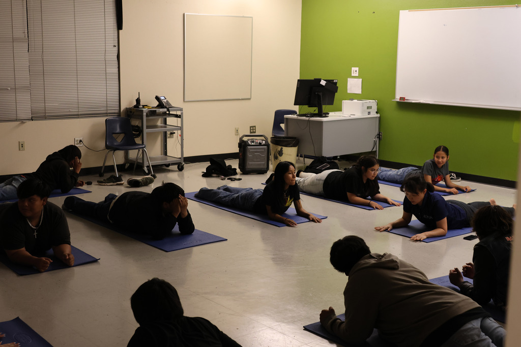 Several individuals are on mats in a room, performing exercises on a floor. Green wall with whiteboard.