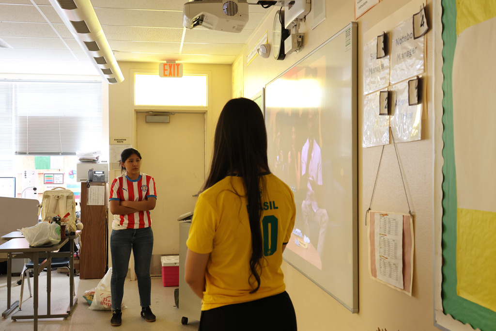 Two individuals in a classroom, one facing a whiteboard, the other with a yellow shirt and a backpack.