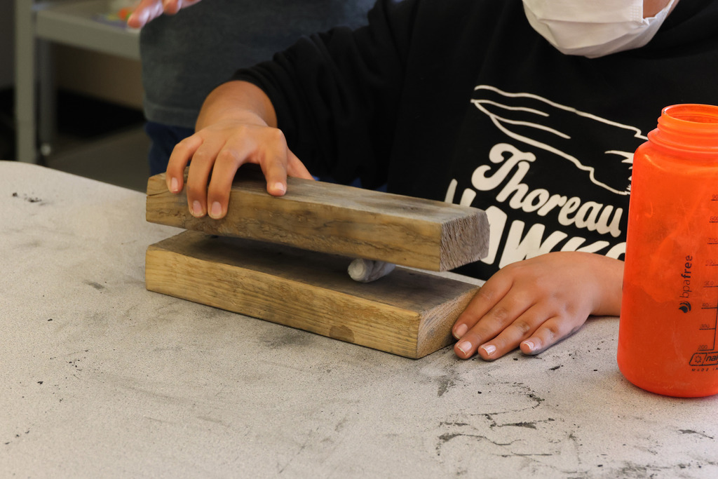 Child wearing a mask uses two wooden boards to crush an object. A bottle is on the right side.