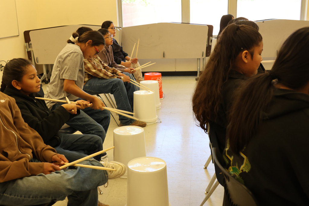 Several people seated in chairs, holding drumsticks, and playing on white drums, with a glass window in the background.