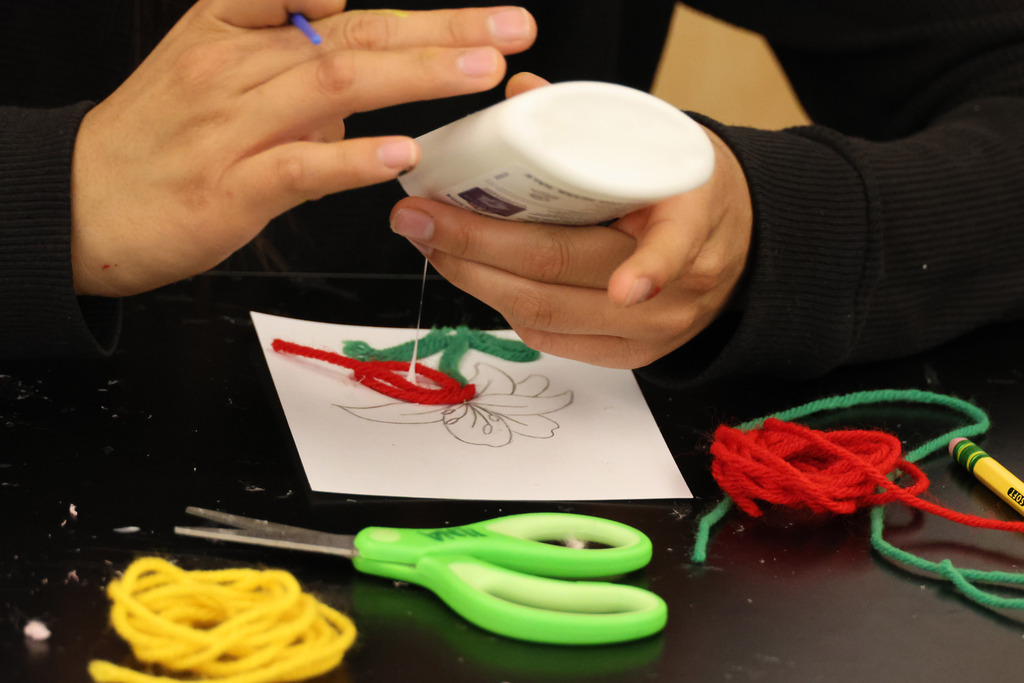 Person using glue and yarn to create a flower on paper. Tools and materials surround the workspace.