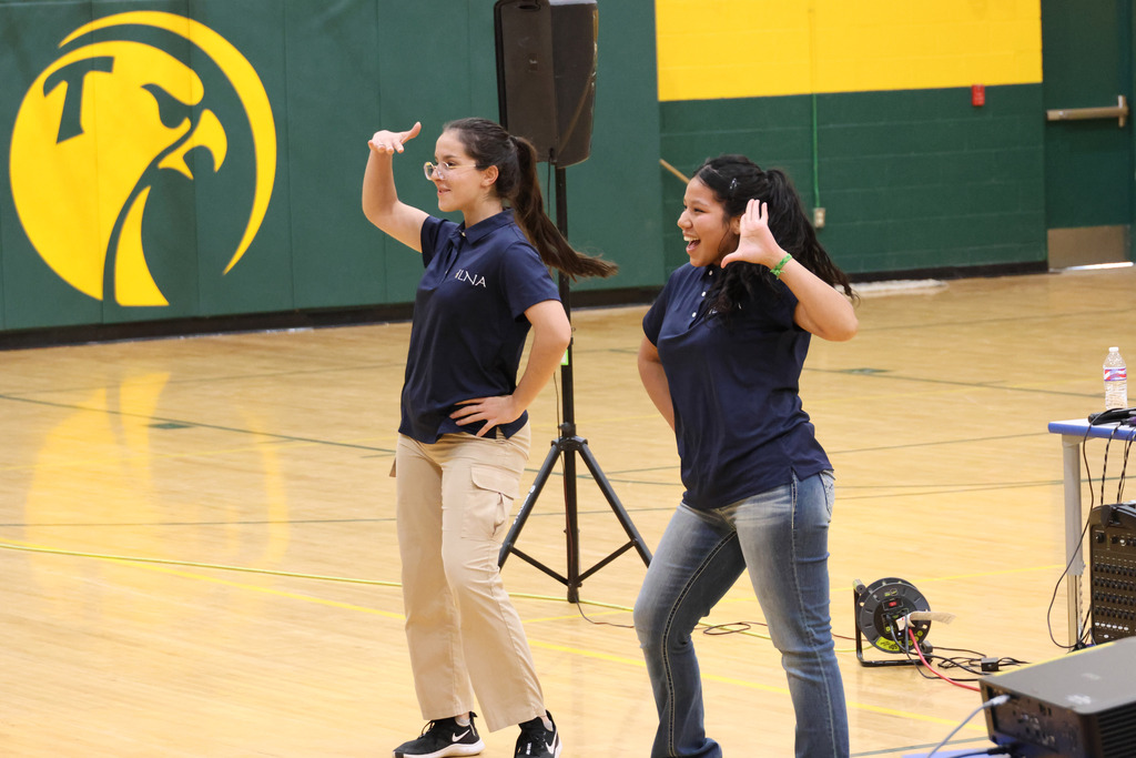 Two women in blue shirts, khakis, and sneakers in a gym with a speaker and a yellow logo.
