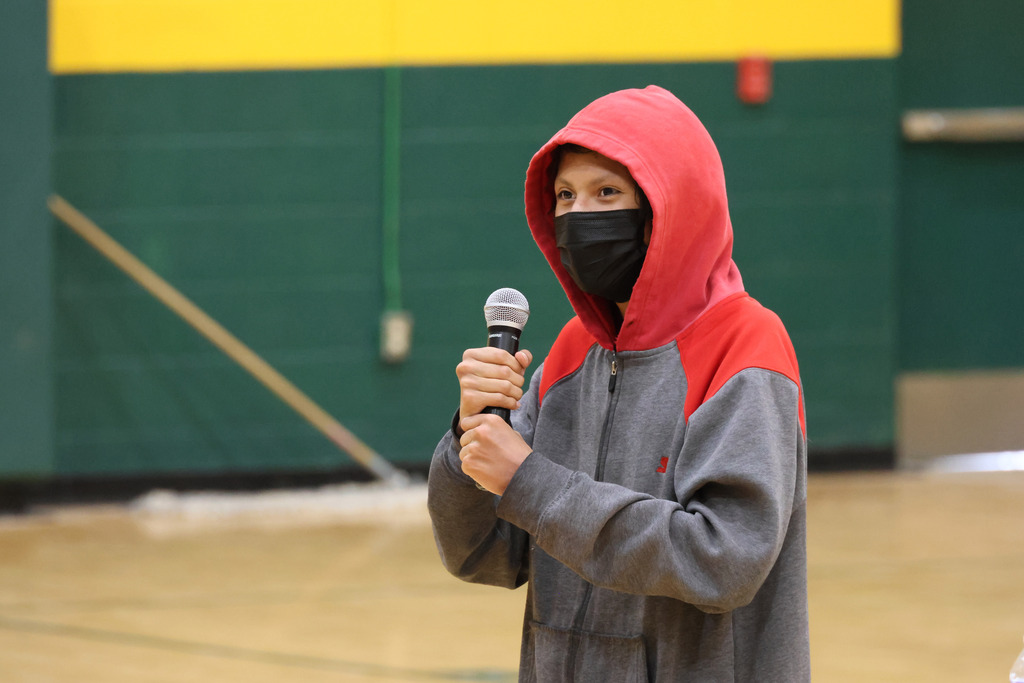 Person in a gray hoodie, red hood, and face mask stands in a gym holding a microphone.