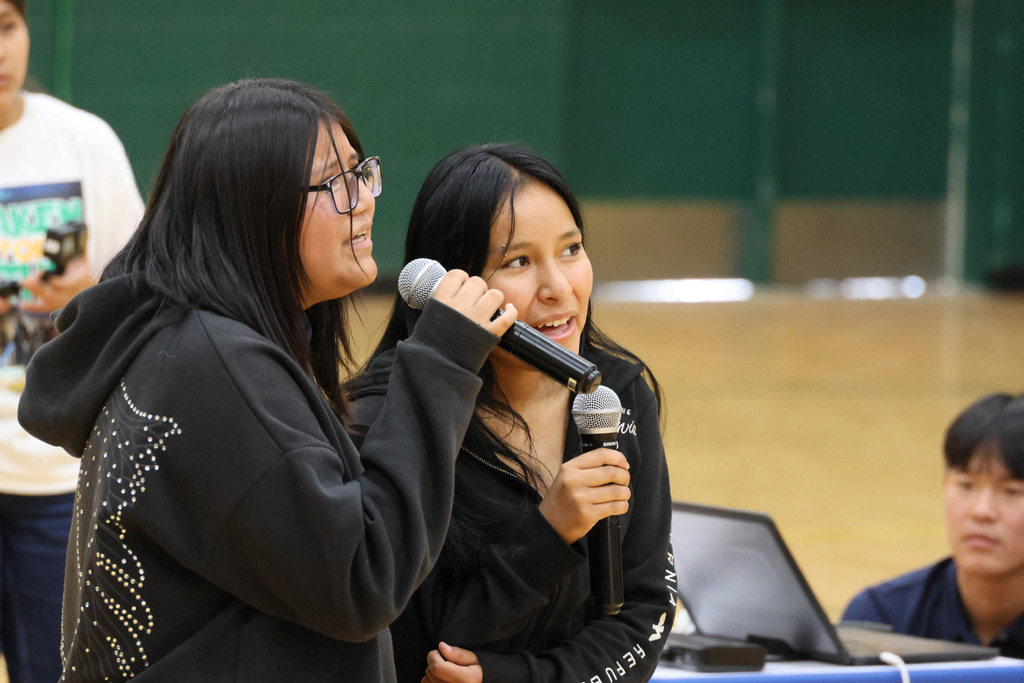 Two women speak into microphones; one wears glasses and a hoodie, the other a black top. Behind them, a laptop rests on a table.