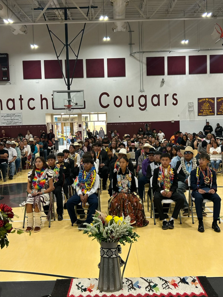Large indoor gathering, people in traditional attire seated in rows, a stage with flowers, and a banner reading "Hatchi Cougars" on the wall.