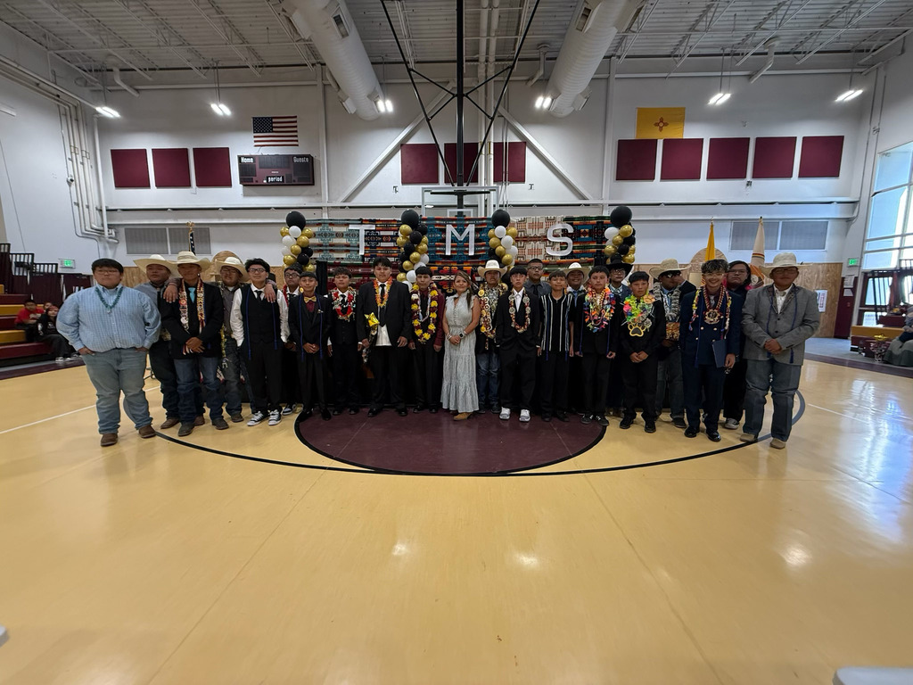 A group of people stands in a gymnasium. They wear formal attire and garlands. The word "TAMS" is displayed on a banner.
