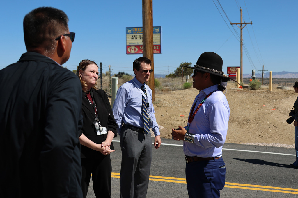 Five individuals in business attire, including a man wearing a hat, converse on a roadside. Background shows desert landscape.