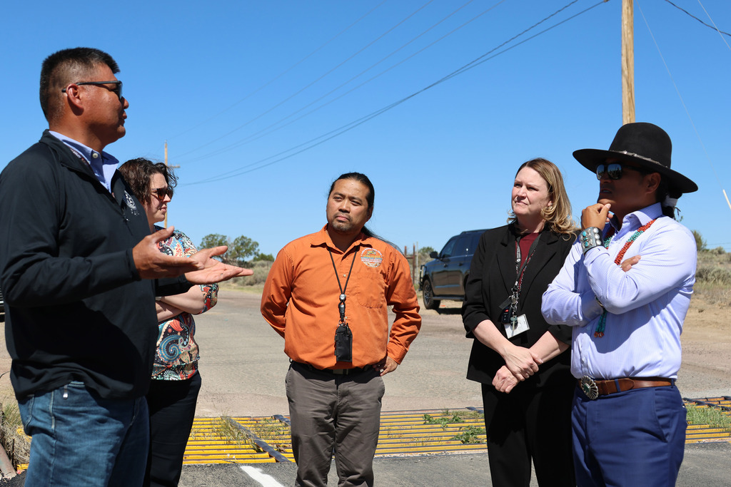 A group of people, including a man in a black jacket gesturing, stands on an empty road with a car visible in the background.