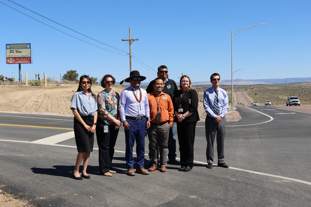 Six people stand on a road with white markings. They wear various clothing. One holds a sign.