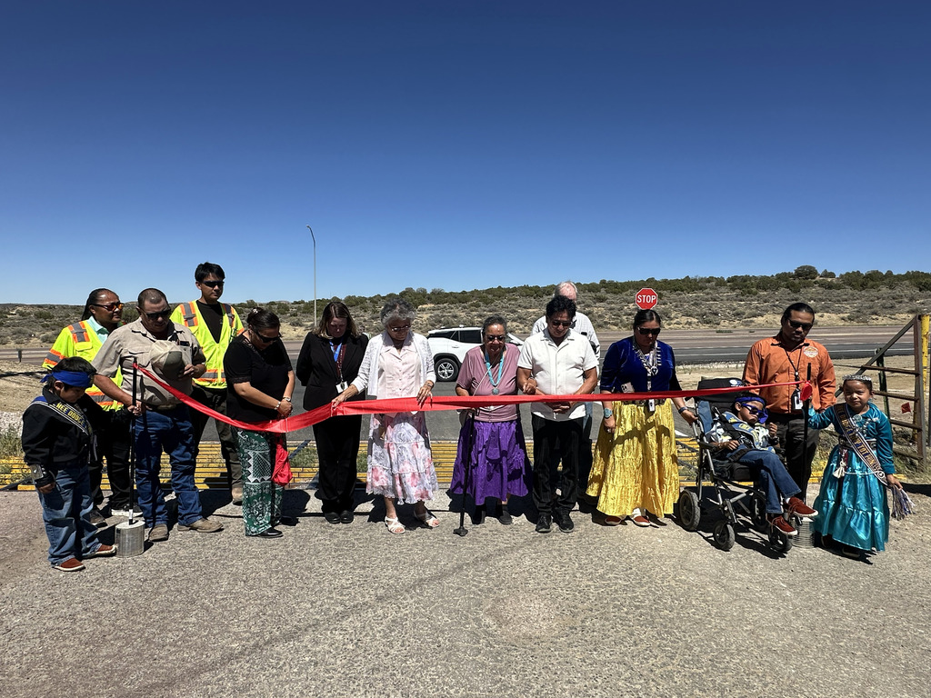 People in traditional clothing stand by a ribbon, holding it up for a photo, with a stop sign and desert in the background.