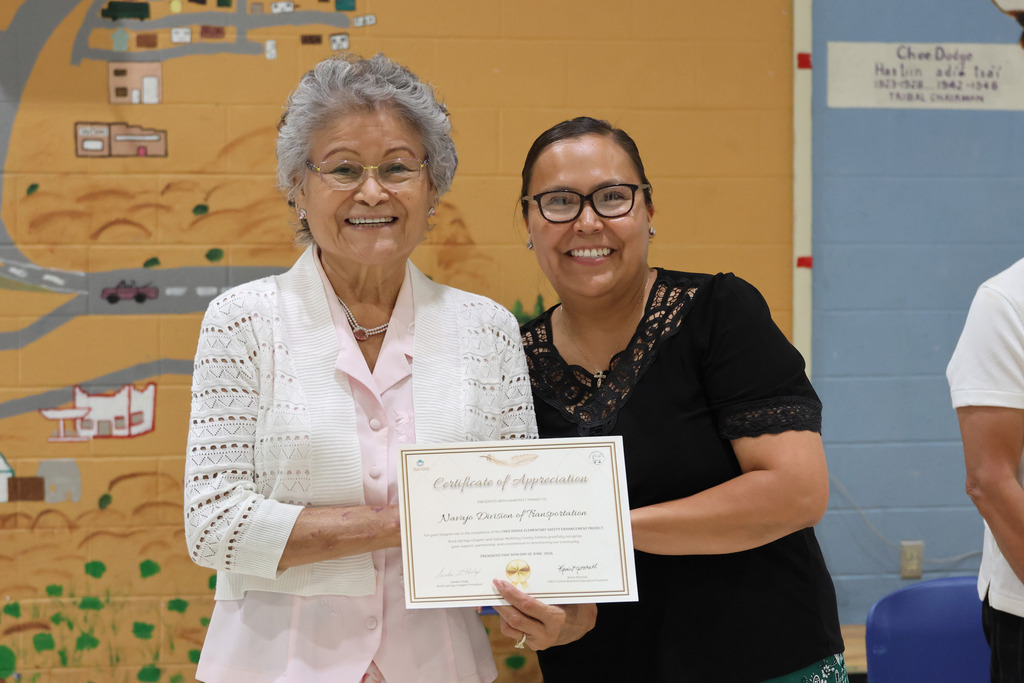 Two women stand next to each other, one holding a certificate. The woman on the left wears a white sweater and glasses. The woman on the right wears glasses and a black top.