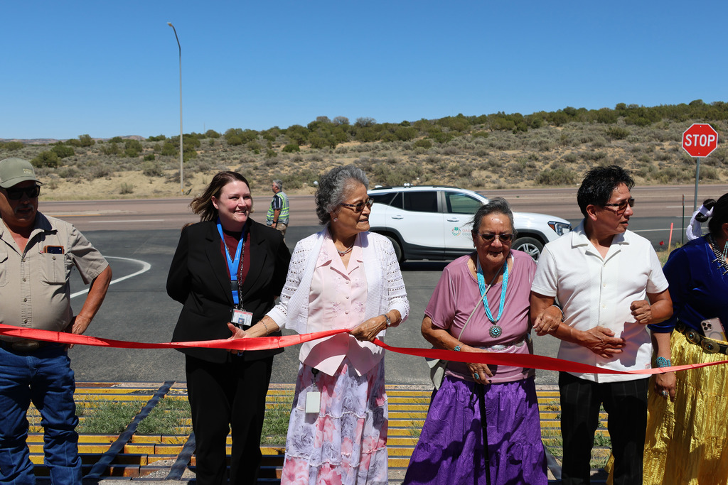 People stand before a red ribbon, holding it. One person cuts it. A car passes in the background.