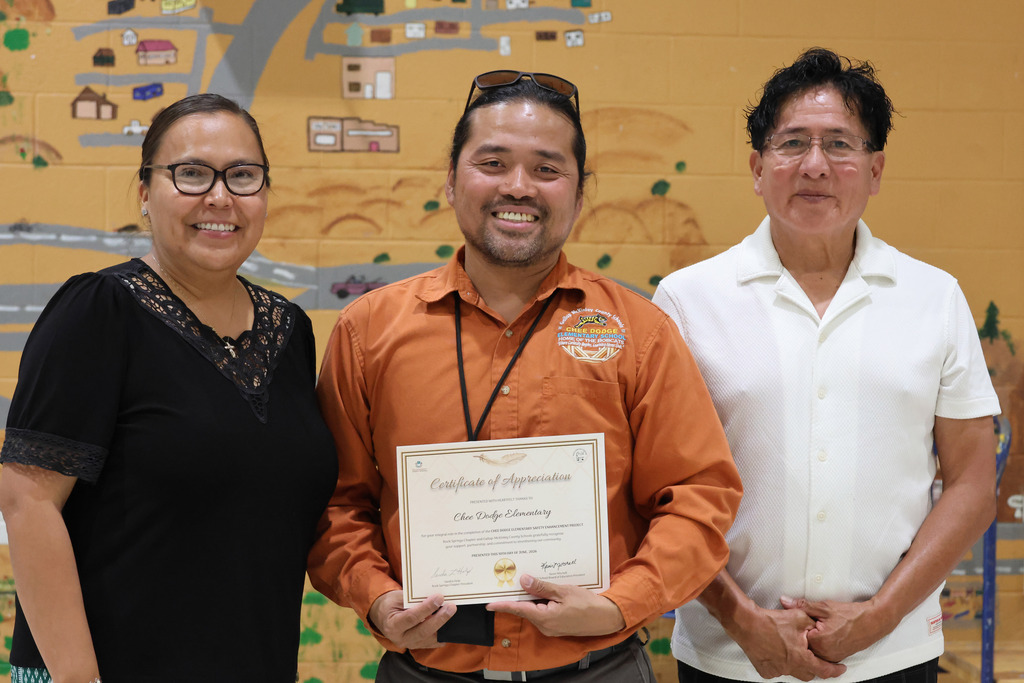 Three adults stand together, smiling. A man holds a certificate. Behind them, a colorful mural depicts a village.