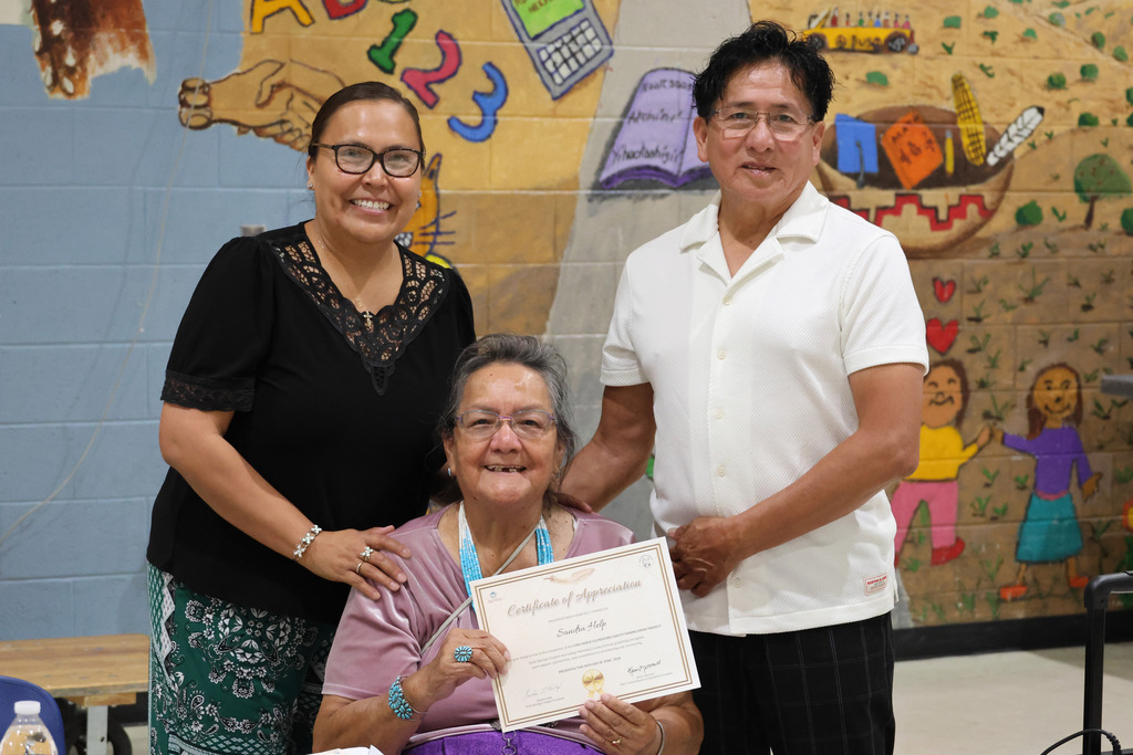 Three people hold a certificate in a classroom with a colorful wall mural. The woman holds a certificate.