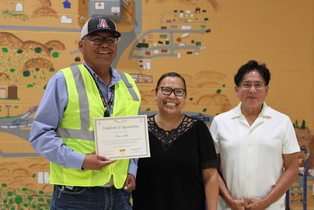 Three individuals stand in front of a mural, with the man on the left holding a certificate.