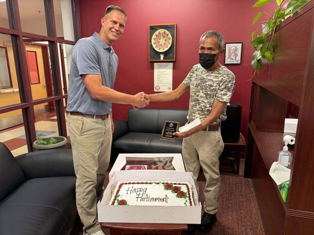 Two men in a room with red walls shake hands. One holds a cake that reads "Happy Retirement".