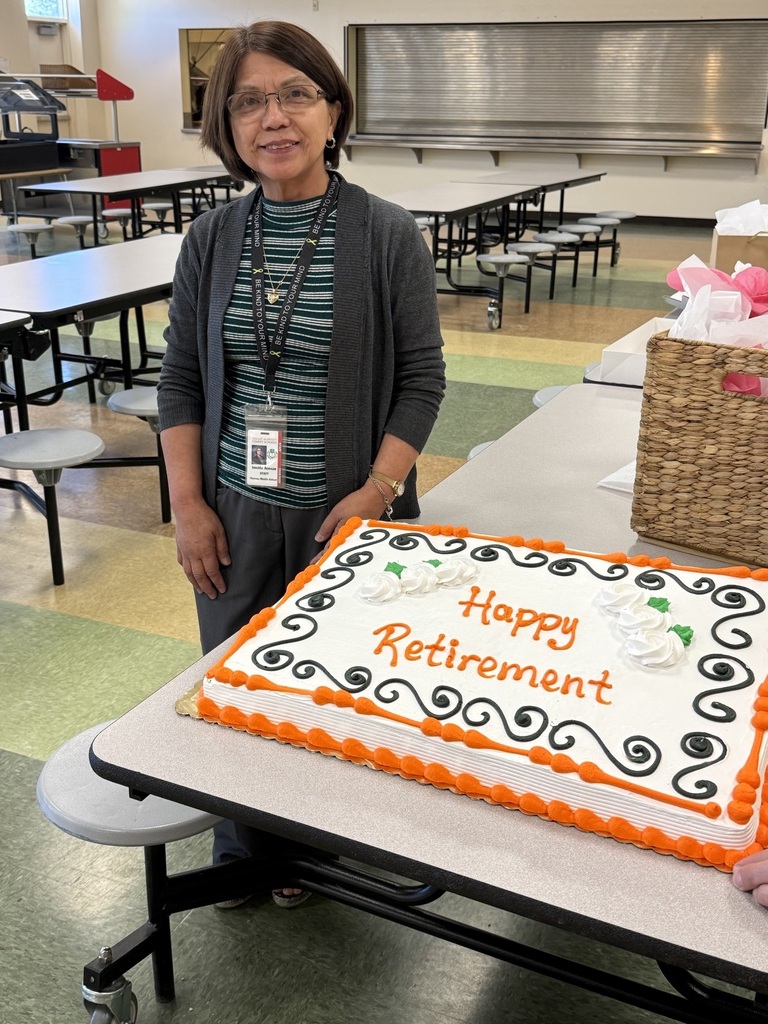 A woman wearing glasses and a lanyard stands next to a cake saying "Happy Retirement" on a table.