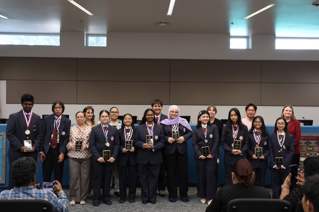 A group of people in suits and ties, possibly students, standing together with medals, in a conference room.
