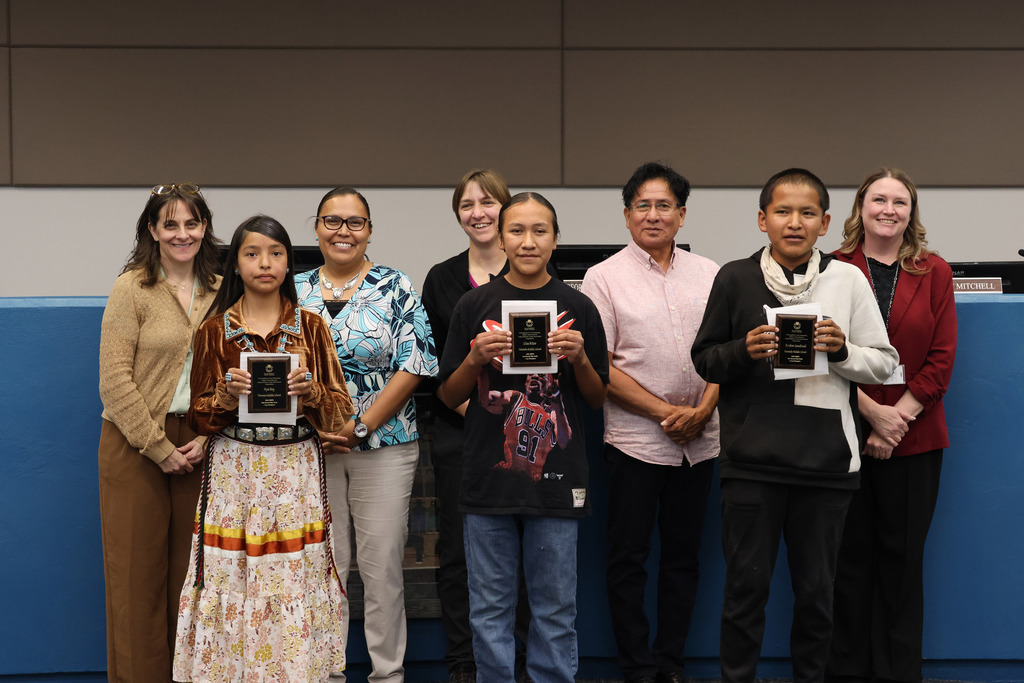 Nine people in two rows hold awards in a room with blue walls. Two women wear dresses and belts.