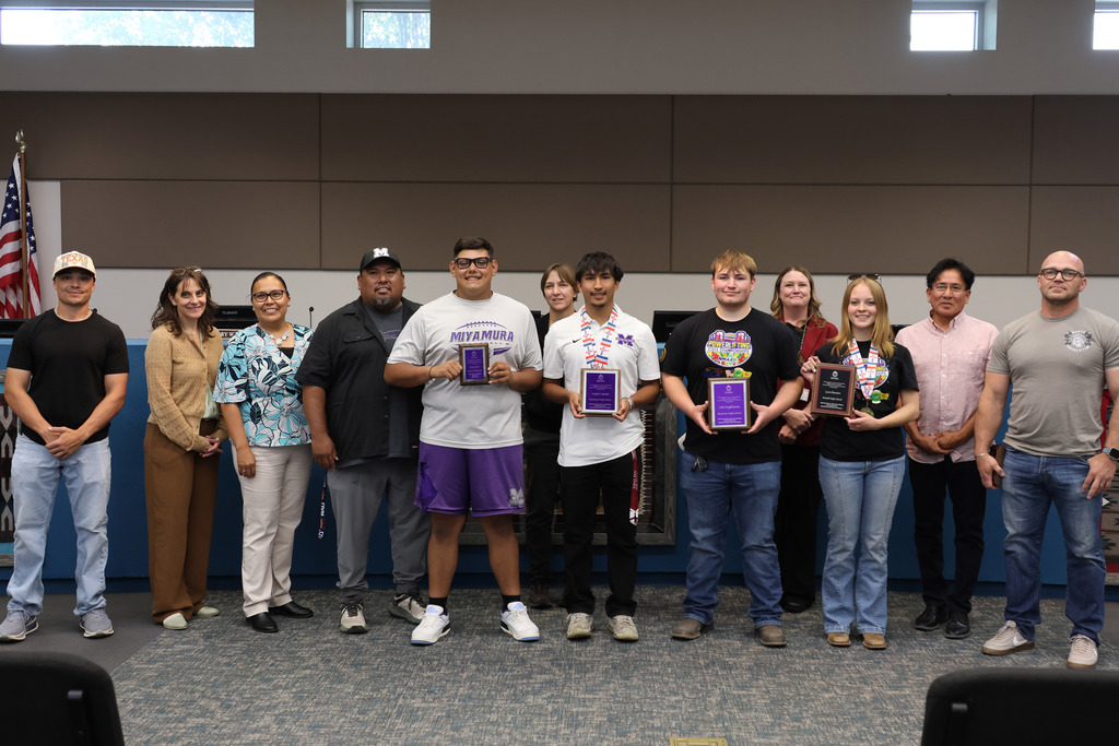 Group of people standing in two rows with awards, one holding a flag in the background.