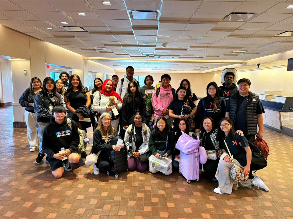 A group of people in an indoor area, possibly a lobby, wearing casual clothing and holding bags.