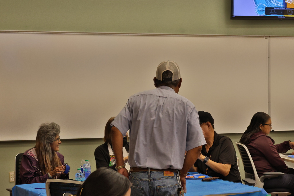 People sitting at a table in a classroom, with a man standing in front of a whiteboard.