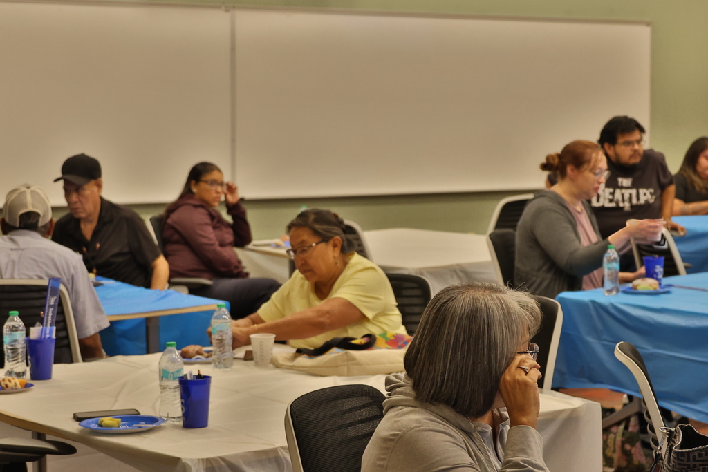 A group of people seated at tables. Blue tablecloths and white table coverings are visible. Water bottles, plates, and napkins on the tables.