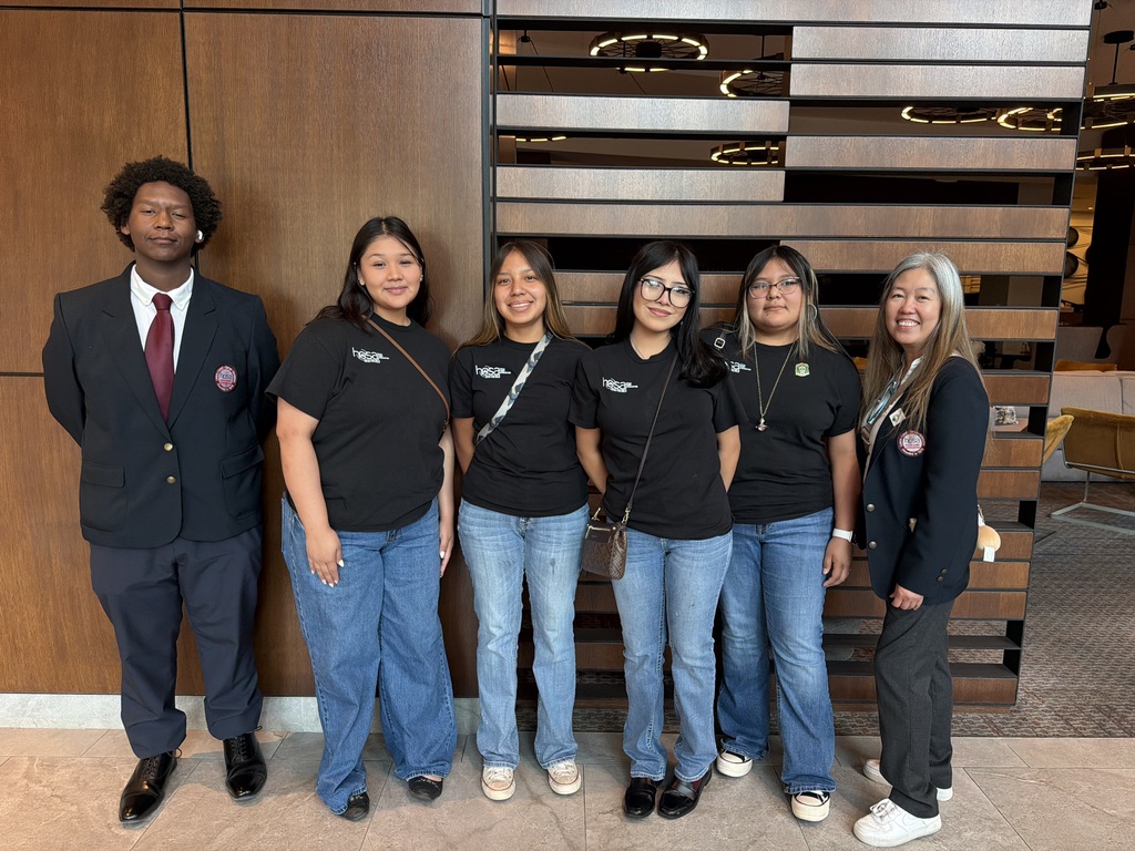 A group of seven individuals dressed in black t-shirts and jeans pose together in front of a wooden paneled wall.