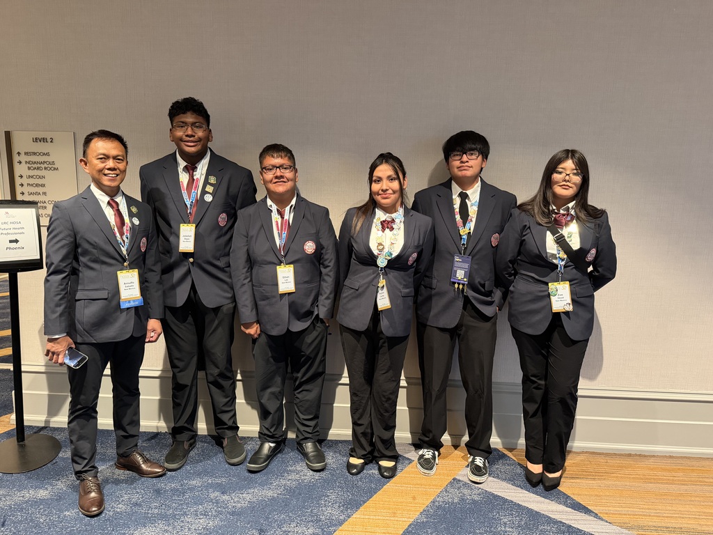 Six individuals in matching blazers, ties, and pins stand against a plain wall. Some wear IDs. Floor has a wood and blue carpet.