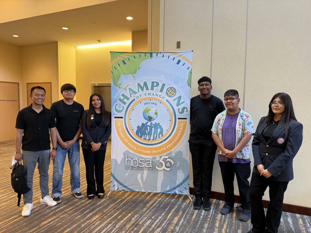 Group of individuals standing in front of a banner with text. They wear casual attire, some carry bags, and pose for a photo.